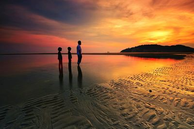 Father with children standing at beach against sky during sunset