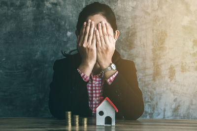 Businesswoman covering face with hands while sitting by coins and model home at table