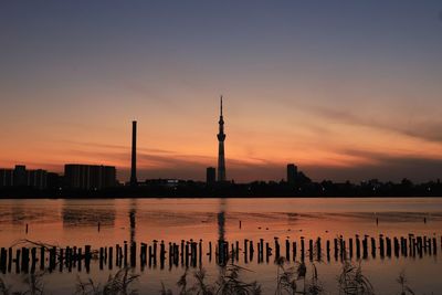 Silhouette buildings by river against sky during sunset