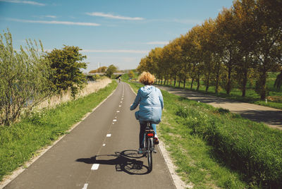 Rear view of woman riding bicycle on road against sky during sunny day