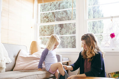 Mother assisting daughter in getting dressed by window at home