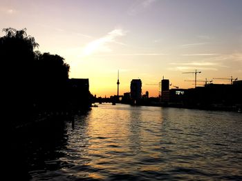 Silhouette buildings by river against sky during sunset