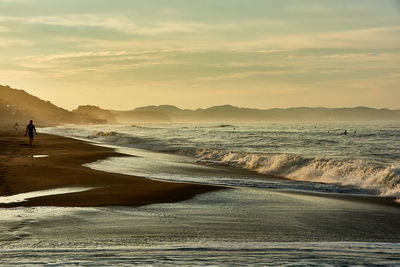 Scenic view of sea against sky with splashing waveform and silhouette people during sunrise