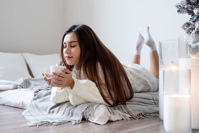 Woman in warm white winter sweater lying in bed at home at christmas eve holding cup