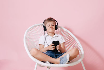 Smiling boy listening music while using phone on chair against colored background