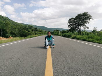 Portrait of man crouching on road against cloudy sky