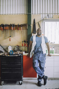 Full length of smiling carpenter leaning on workbench in workshop