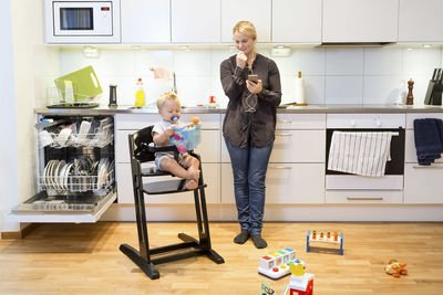 Mother talking on mobile phone in kitchen while baby boy sitting on high chair
