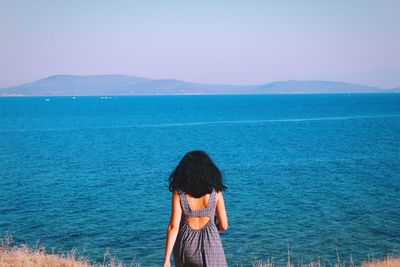 Rear view of woman standing of field by sea against clear sky