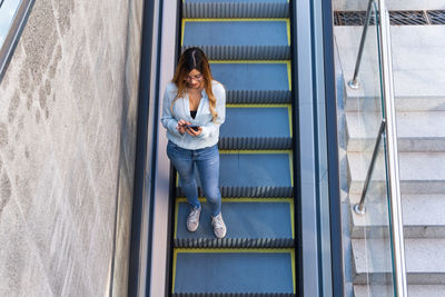 Cheerful lady with smartphone walking down on the escalator while smiling, photo from above