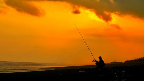 Silhouette man with fishing rod at beach against orange sky