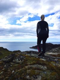 Rear view of woman looking at sea while standing on rock against cloudy sky