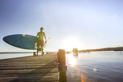 Shirtless man walking with paddleboard on pier over lake against sky