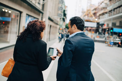 Rear view of businesswoman showing digital tablet to businessman while walking on sidewalk in city