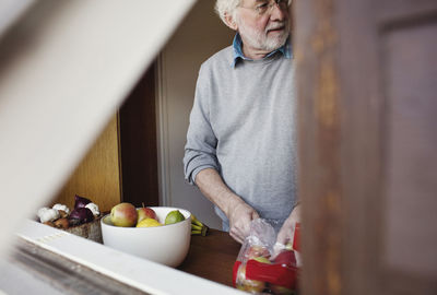 Senior man removing apples from packet by container while standing at kitchen counter