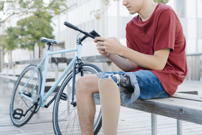 Teen male with leg prosthesis sitting on bench and browsing smartphone