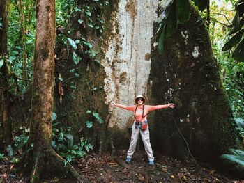 Happy woman with arms outstretched standing against huge tree at forest