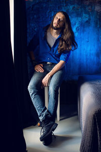 Man with long hair and beard sits at home on a chair by the window in a blue shirt and jeans
