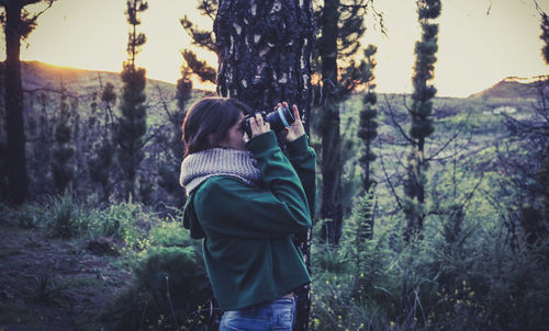 Side view of woman looking through binoculars against trees at forest