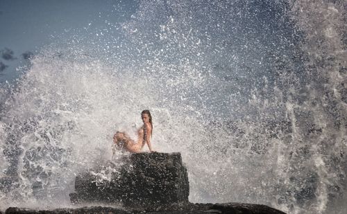 Naked woman sitting on rock by wave splashing in sea