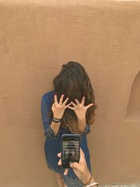 Cropped image of hand photographing young female friend while hiding face against wall