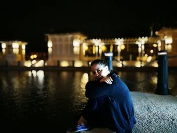 Portrait of mature woman sitting by lake against illuminated building at night