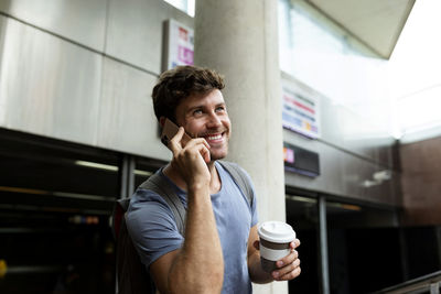 Smiling young commuter talking on smart phone while looking away and holding reusable coffee cup at subway station