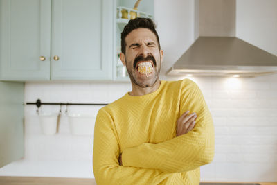 Man with tangerine fruit in mouth and arms crossed at kitchen