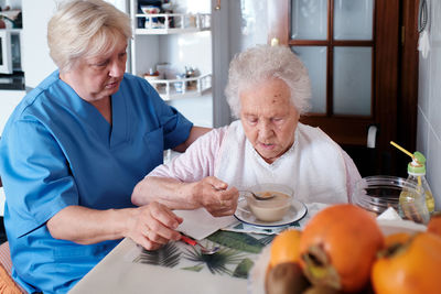 Nurse in uniform sitting near aged woman in wheelchair eating tasty soup at table with food in light kitchen at home