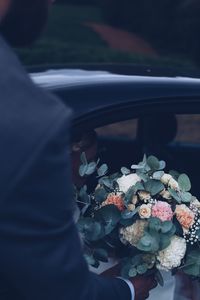 Cropped hand of bridegroom holding bouquet by car