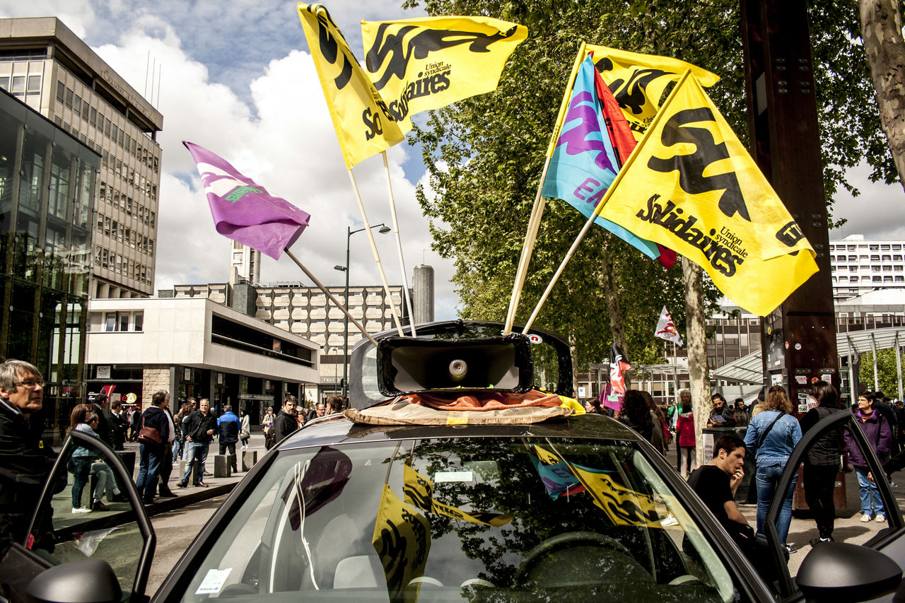 car, mode of transportation, transportation, vehicle, city, motor vehicle, architecture, group of people, crowd, land vehicle, large group of people, flag, street, building exterior, built structure, text, adult, men, day, protest, outdoors, women, communication, sign, nature, person, travel, road