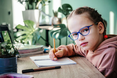 Primary school girl in eyeglasses writing in her notebook while having online lesson during covid