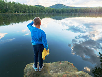 Blue wearing boy with  towel and swimming googles in hands stay on jumping cliff above lake water