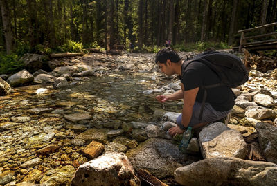 Brown indian man with backpack sitting next to clean water stream or river drinking water from it