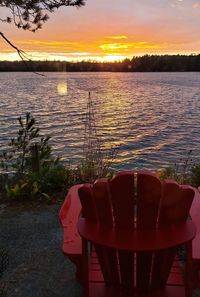 Empty chairs and table by lake against sky during sunset