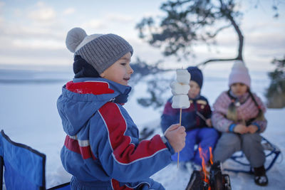 Happy friends sit around campfire on shore of frozen lake in forest and fry marshmallows