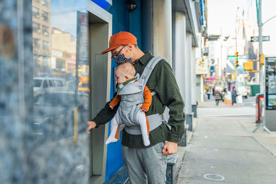 Father with baby daughter using atm at sidewalk in city during covid-19