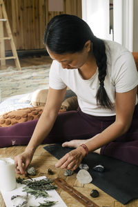 Young woman with crystals and herbs sitting on exercise mat at home