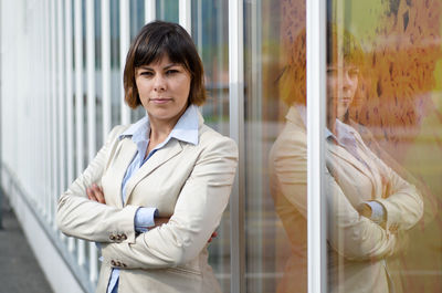 Portrait of confident businesswoman with arms crossed standing by glass door
