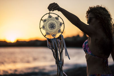 Woman holding dreamcatcher at beach against sky during sunset