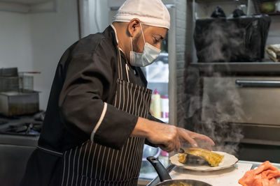 Side view of mature ethnic male cook in sterile mask with spatula putting hot pasta on plate in restaurant kitchen