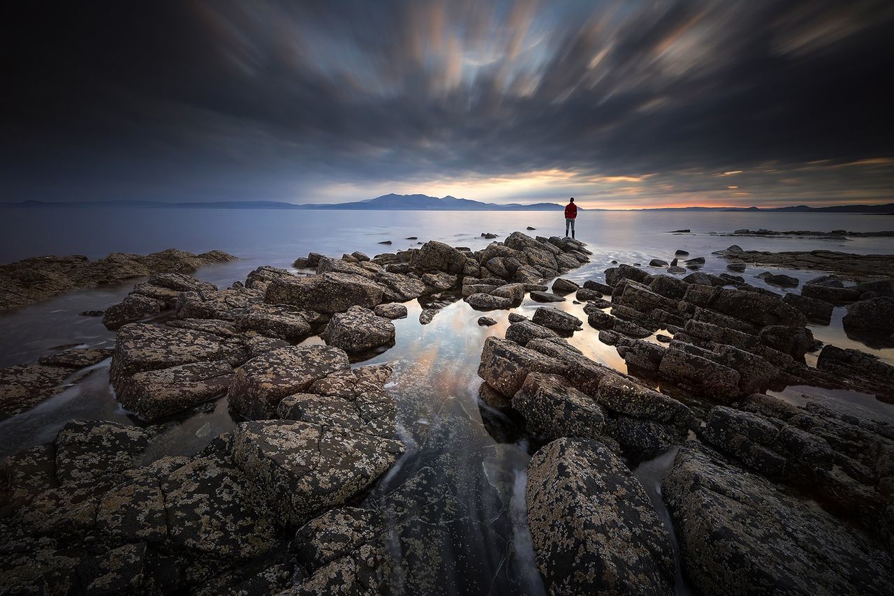 Rear view of man standing on rock at beach against cloudy sky