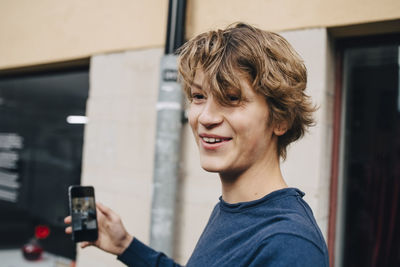 Close-up of smiling teenage boy with mobile phone by building