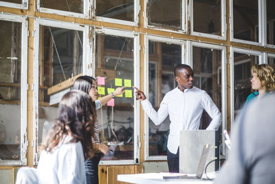 Serious creative professionals pointing at adhesive notes while discussing with team in office