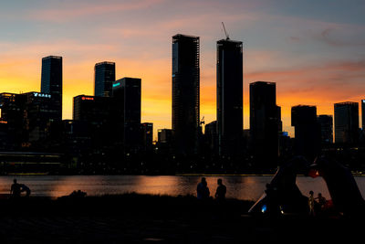 Modern buildings by river against sky during sunset