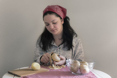 Young woman peeling potatoes while sitting at table against wall