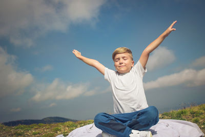 Cute boy sitting with arms outstretched on rock against sky