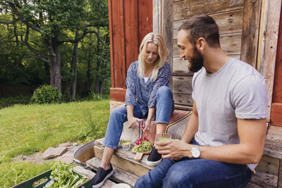 Man having drink looking at friend cutting vegetables outside cottage