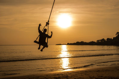 Silhouette person swinging against calm sea at sunset