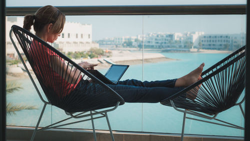 Full length of woman working on laptop while sitting on chair in balcony against lake